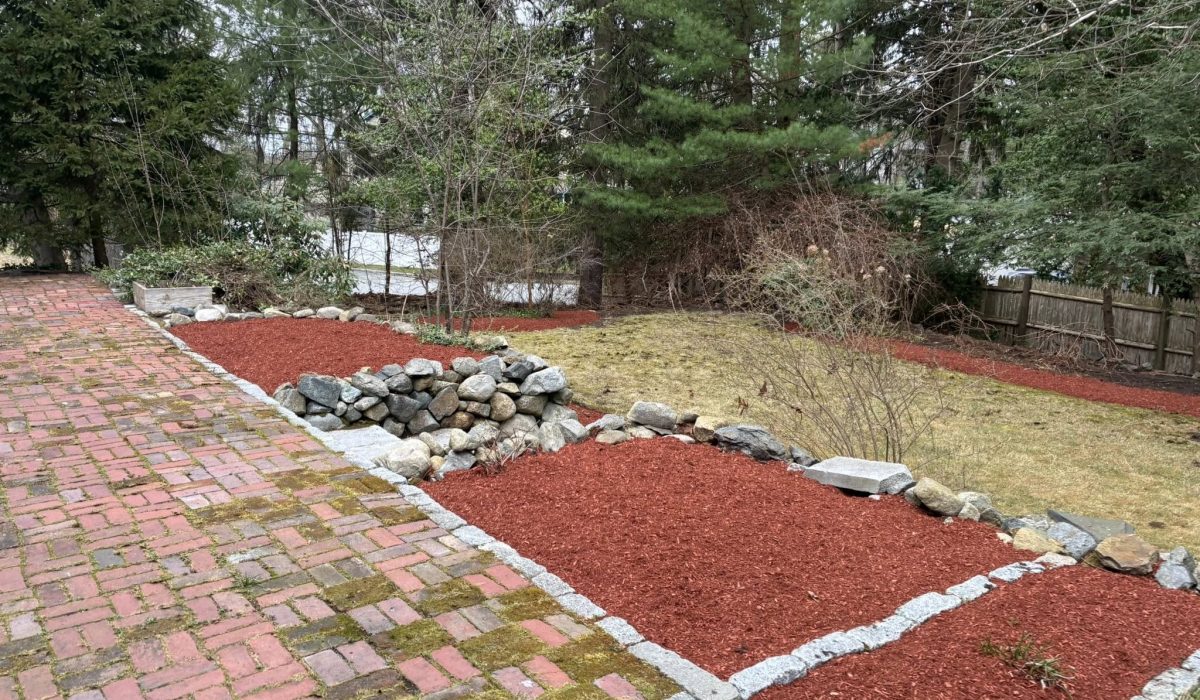 Fresh red mulch landscape bed installation with granite edging and decorative stone borders along a brick driveway.