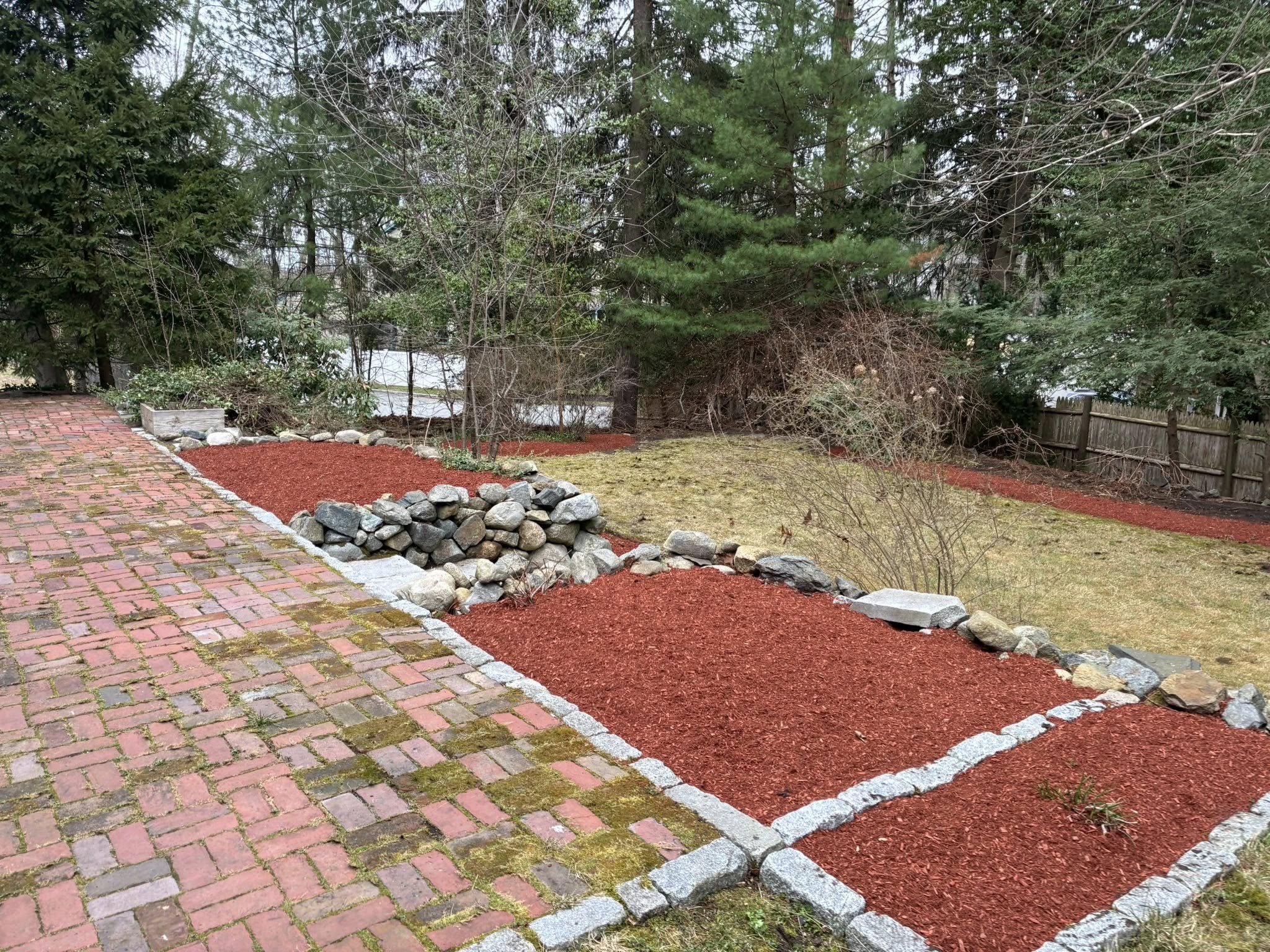 Fresh red mulch landscape bed installation with granite edging and decorative stone borders along a brick driveway.